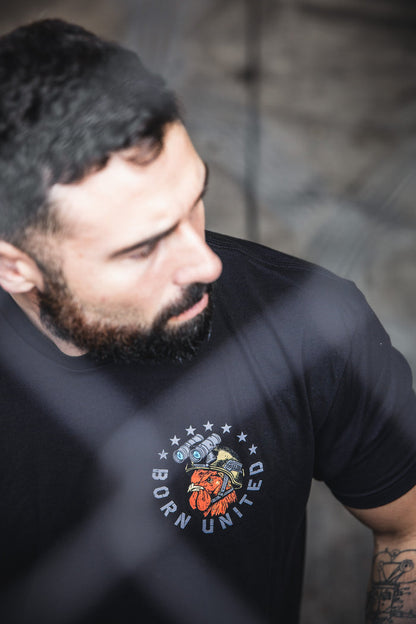 A bearded man with short dark hair wears a black Battle Bird t-shirt by Born United Private Label, made from a 60/40 blend and featuring a heart, camera, and stars graphic. The background is neutral and out of focus.