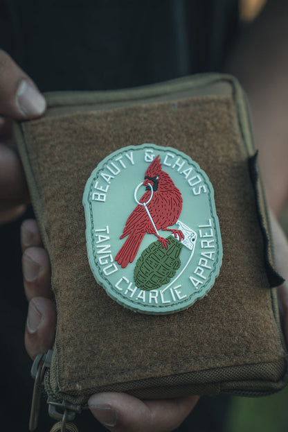 Two hands hold a durable PVC patch showcasing a red cardinal on a green grenade. The text Beauty & Chaos is displayed above, with Tango Charlie Apparel below, set against a softly blurred background that highlights the intricate design of this unique Beauty and Chaos 2.0 patch.