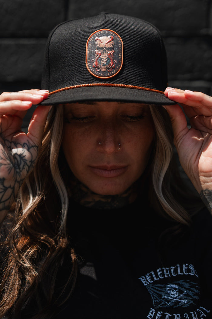 A tattooed individual adjusts the Between The Devil & Deep Blue Sea Snapback by Relentless Betrayal while outdoors. The black hat showcases an embroidered skull patch for a bold, edgy style. They also have a nose piercing and are dressed in a black shirt.