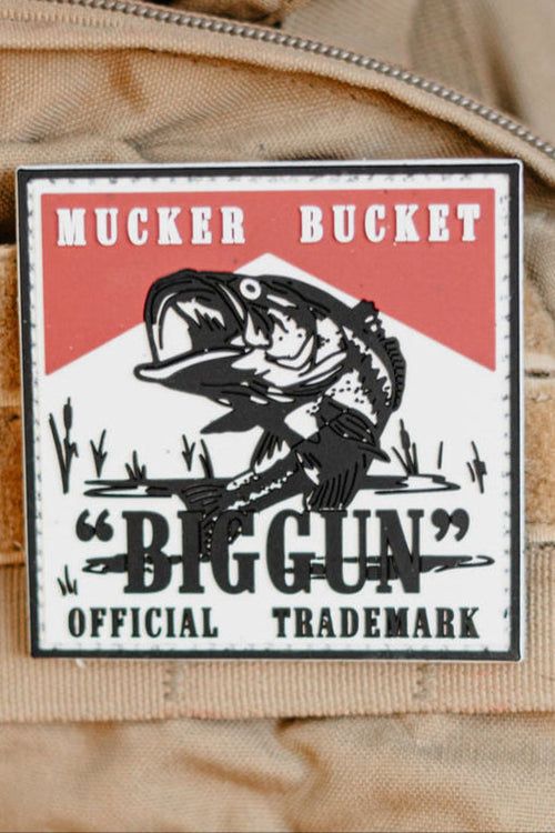 The "Biggun" PVC patch by Mucker Bucket displays a fish design and the text: Mucker Bucket Big Gun Official Trademark on tan fabric with Velcro backing.