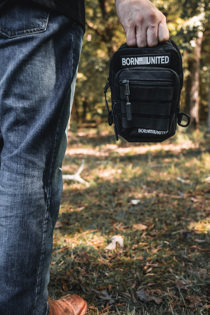 In a wooded area, only the lower body and hand of a person are visible as they hold the Black Phantom Tactical Sling-Pac - 4L by Born United Private Label. They stand on grass, wearing blue jeans and brown shoes.