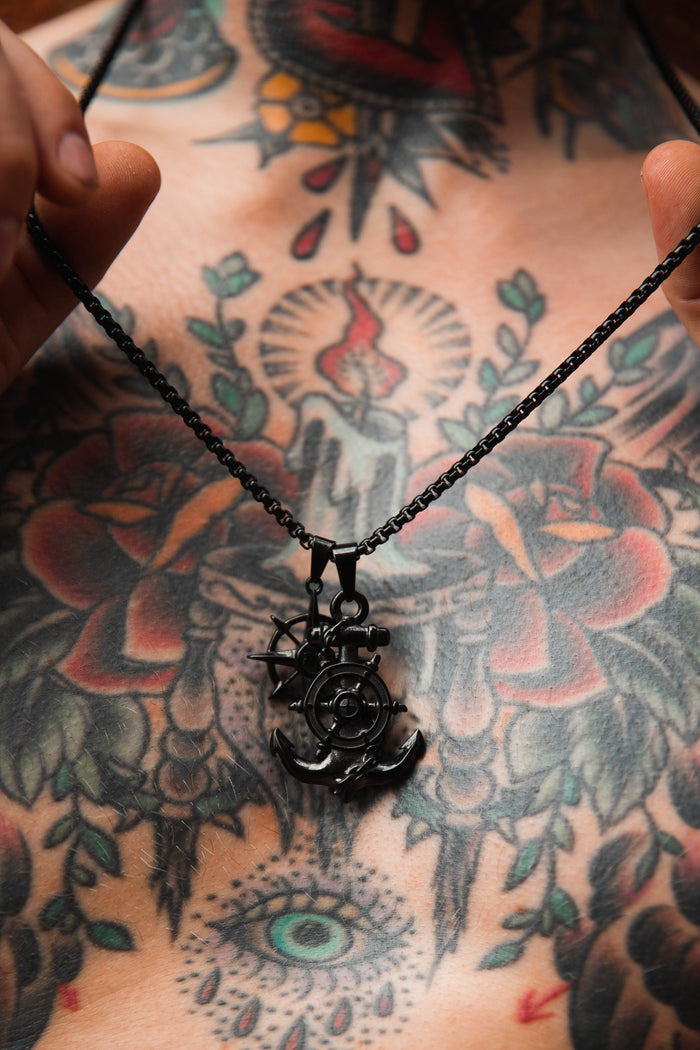 A close-up shows a person holding the Relentless Betrayal Blackout Anchors Aweigh Necklace against a tattooed chest adorned with colorful roses, an eye, and abstract designs; the pendant hangs from a durable stainless steel chain built to last.