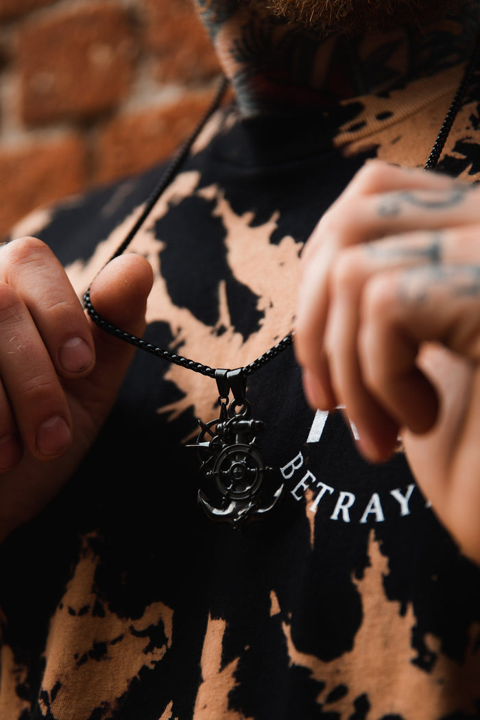 A person in a tie-dye shirt holds the Relentless Betrayal Blackout Anchors Aweigh Necklace, featuring a handcrafted metal pendant on a sleek stainless steel chain. Text on the shirt is partially visible against a blurred brick wall background.