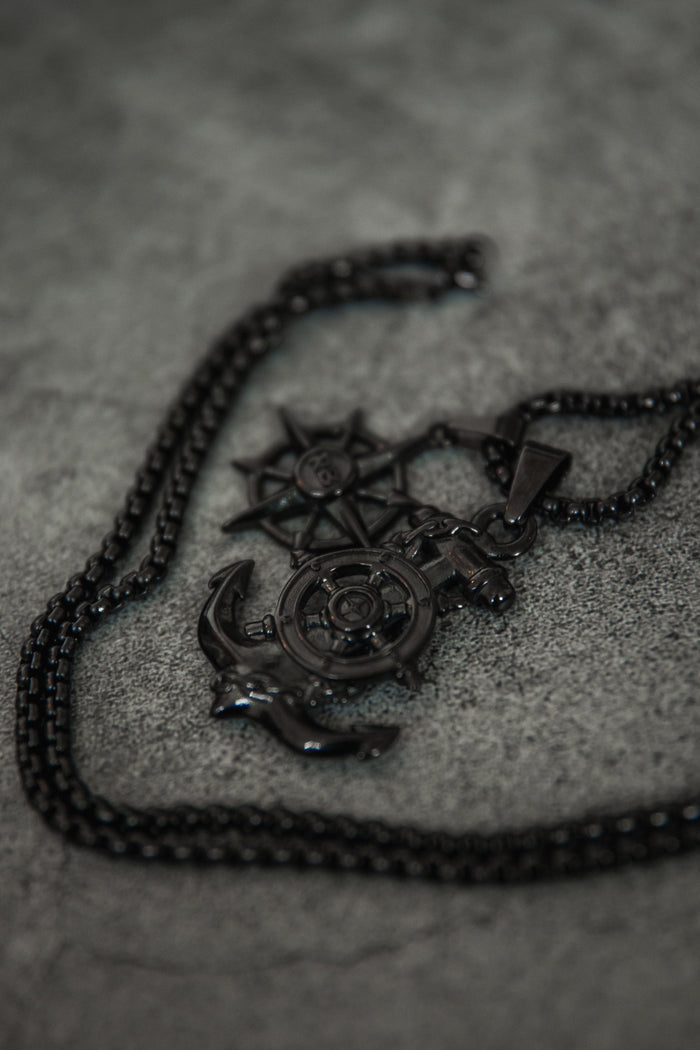The Blackout Anchors Aweigh Necklace by Relentless Betrayal features nautical-themed pendants like an anchor, ship wheel, and compass star on a handcrafted stainless steel chain. It includes a lifetime warranty for enduring elegance against a textured gray background.