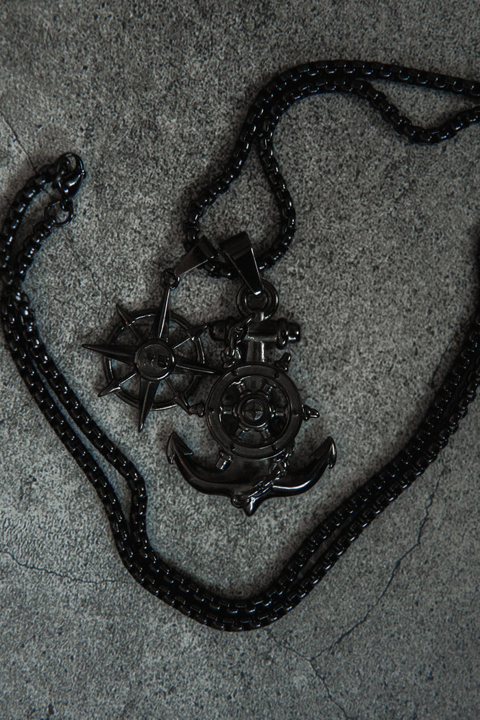 The Relentless Betrayal Blackout Anchors Aweigh Necklace rests on a textured gray surface, showcasing a handcrafted ships wheel, anchor, and compass rose pendant. Its durable stainless steel chain includes a lifetime warranty for peace of mind.