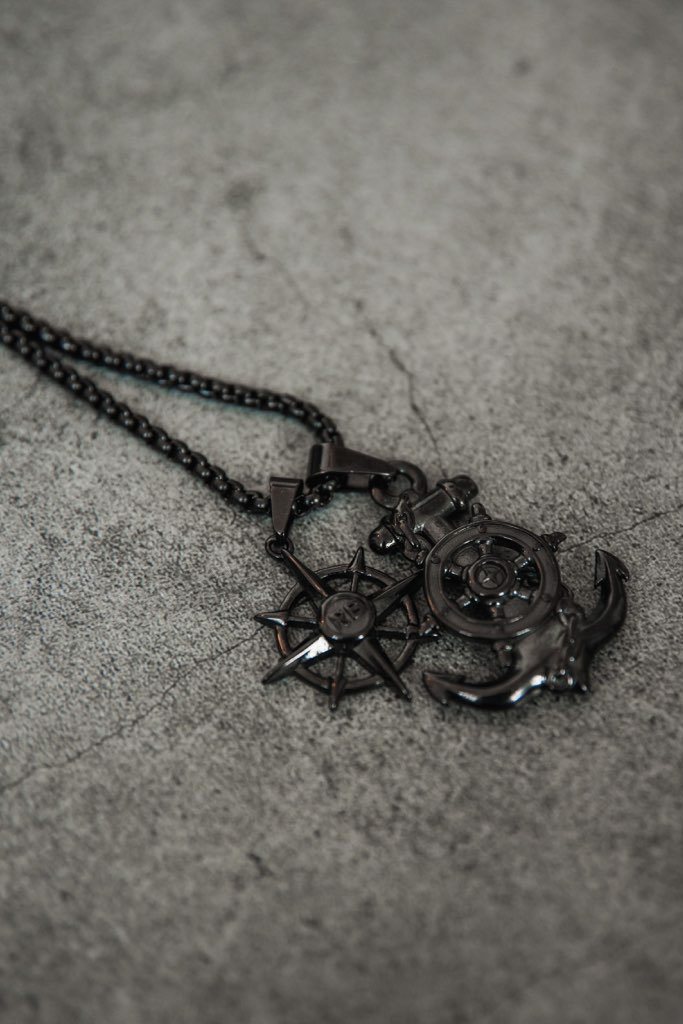 The Relentless Betrayal Blackout Anchors Aweigh Necklace is a handcrafted black stainless steel chain featuring two pendants: an intricate anchor with rope and crossbones, and a detailed compass rose, all on a textured gray surface. It includes a lifetime warranty for peace of mind.
