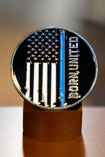 The Blue Line Flag Challenge Coin by Born United Private Label features a black-and-white American flag with a blue stripe and "Born United" along the right, displayed upright on a brown stand.