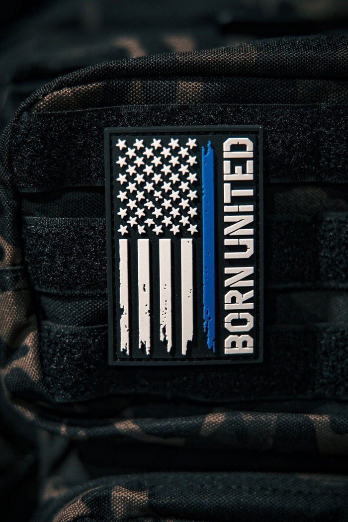A Blue Line Flag PVC Patch by Born United Private Label, featuring a black, white, and blue American flag with a single blue stripe and BORN UNITED text, is attached to a black tactical backpack.