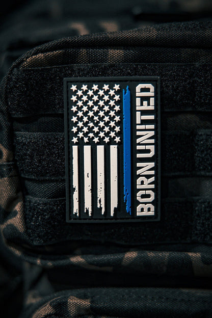 A Blue Line Flag PVC Patch by Born United Private Label, featuring a black, white, and blue American flag with a single blue stripe and BORN UNITED text, is attached to a black tactical backpack.