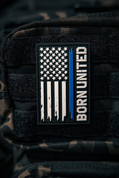 A Blue Line Flag PVC Patch by Born United Private Label, featuring a black, white, and blue American flag with a single blue stripe and BORN UNITED text, is attached to a black tactical backpack.