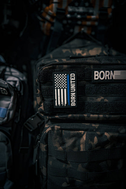 A camouflage backpack displays the Born United Private Label Blue Line Flag PVC Patch featuring a black-and-white American flag with a blue stripe. More backpacks and gear appear in the background.