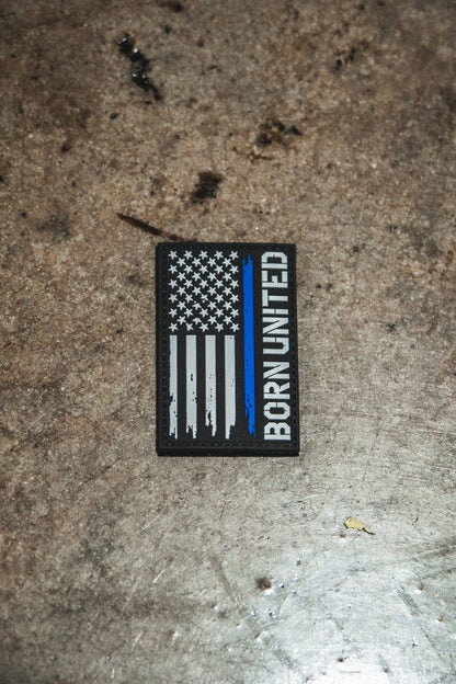 The Blue Line Flag PVC Patch by Born United Private Label features a black, white, and blue American flag with BORN UNITED written vertically on the right side, displayed on a textured surface.