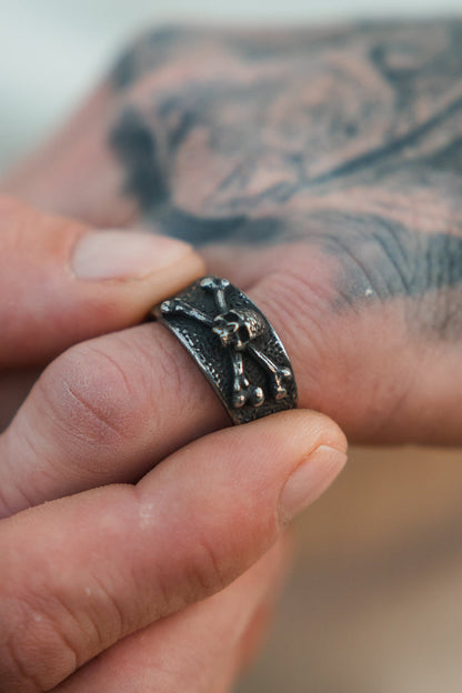 A close-up shows a hand wearing the Relentless Betrayal Bone Crusher Ring, crafted in stainless steel with a raised skeleton design. A tattoo is partially visible on the finger holding the ring.
