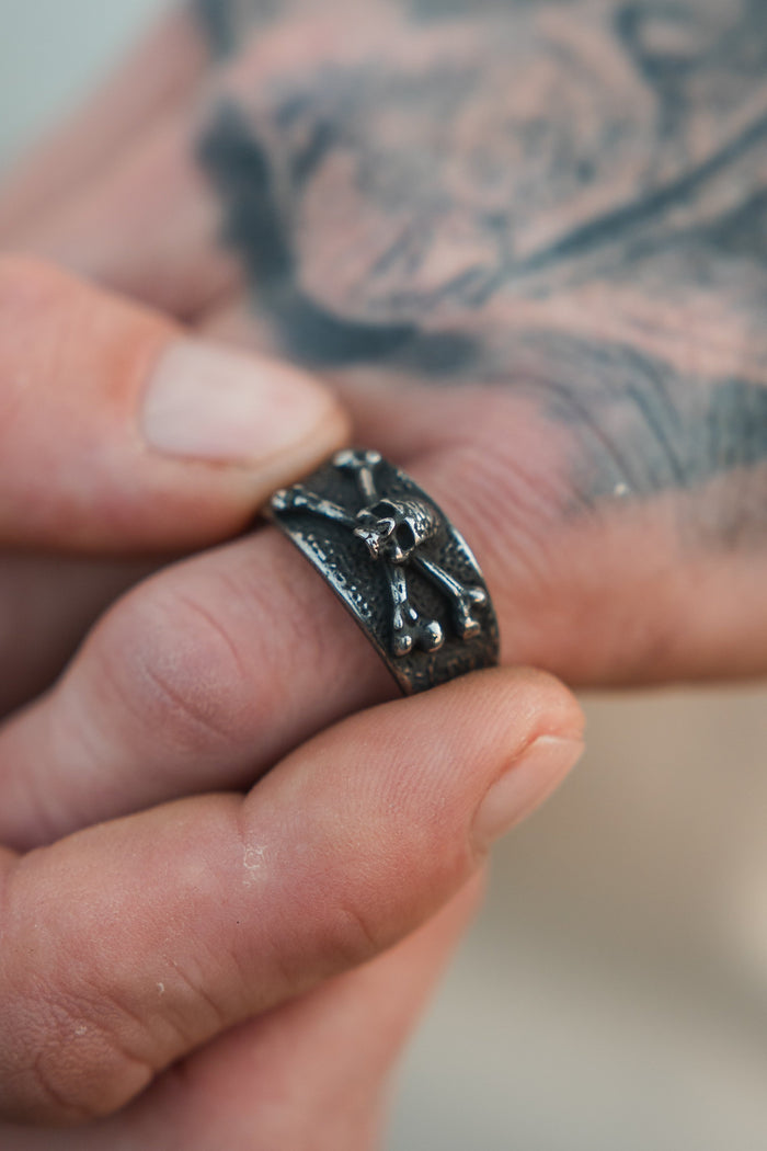 A tattooed hand holds the Relentless Betrayal Bone Crusher Ring, a stainless steel piece adorned with raised animal and bone details.