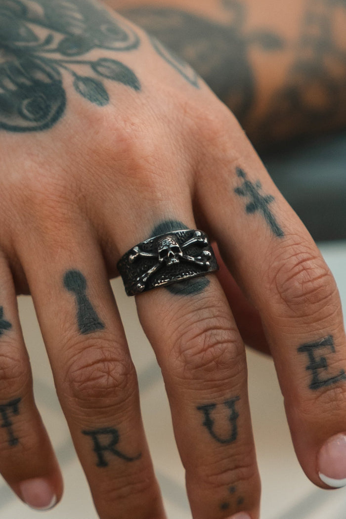 A close-up of a tattooed hand with "TRUE" on the fingers, wearing the Relentless Betrayal Bone Crusher Ring—a textured black ring with a skull design, creating a bold pirate-inspired look.