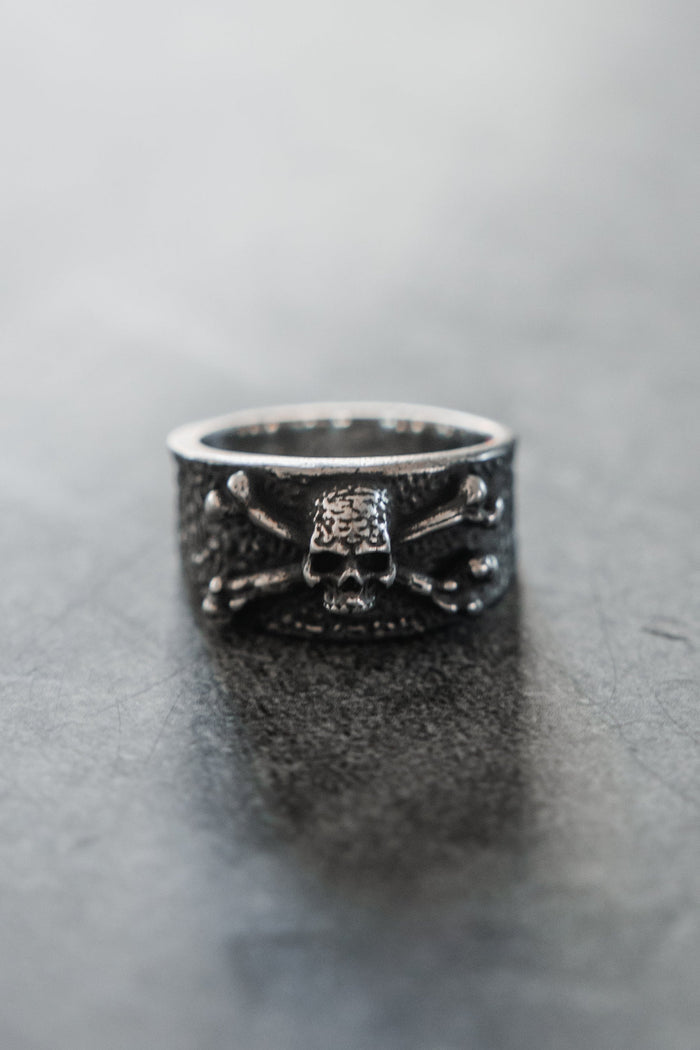 The Relentless Betrayal Bone Crusher Ring, a silver piece with a raised skull and crossbones, stands upright on a smooth gray surface. The softly blurred background highlights the bold details of this striking ring.
