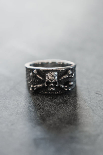 The Relentless Betrayal Bone Crusher Ring, a silver piece with a raised skull and crossbones, stands upright on a smooth gray surface. The softly blurred background highlights the bold details of this striking ring.