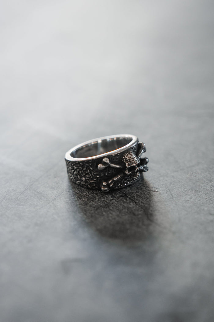 The Bone Crusher Ring by Relentless Betrayal features a textured silver band with a skull and crossbones design, displayed on a smooth gray surface—ideal for fans of pirate rings.