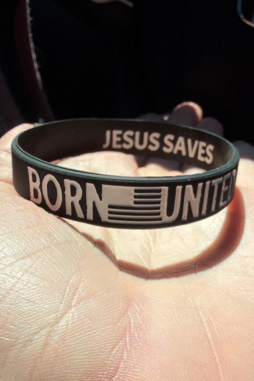 A hand displays the Born United Bracelet by Born United Private Label—a black silicone wristband with "BORN UNITED" and a flag on the outside, "JESUS SAVES" inside—illuminated by sunlight on the person’s palm.