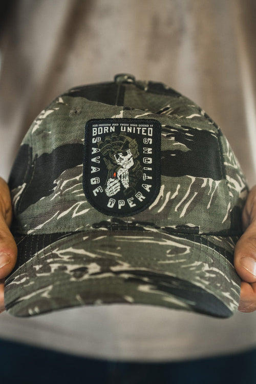 A person holds the Born United Buckle Hat - Savage Operations by Born United Private Label, a camouflage cap featuring a patch with "Born United Savage Operations" and an image of a skeleton holding a rifle.