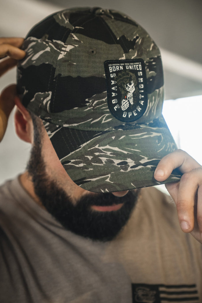 A bearded man in a camouflage Born United Private Label Buckle Hat – Savage Operations, holds the brim and partly obscures his face.