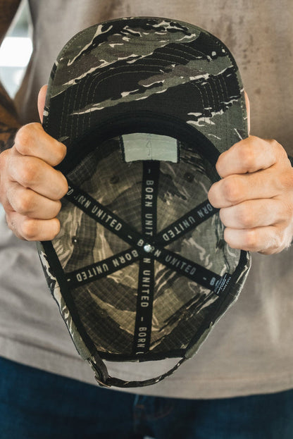 A tattooed individual holds the Born United Buckle Hat - Savage Operations by Born United Private Label, displaying its interior black straps labeled BORN UNITED. They wear a gray shirt, blue jeans, and a black wristwatch.