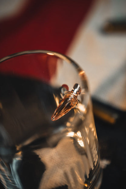 A close-up of the Born United Bullet Whiskey Glass by Born United Private Label, showcasing a bullet embedded in its side against a blurred red, white, and black backdrop.