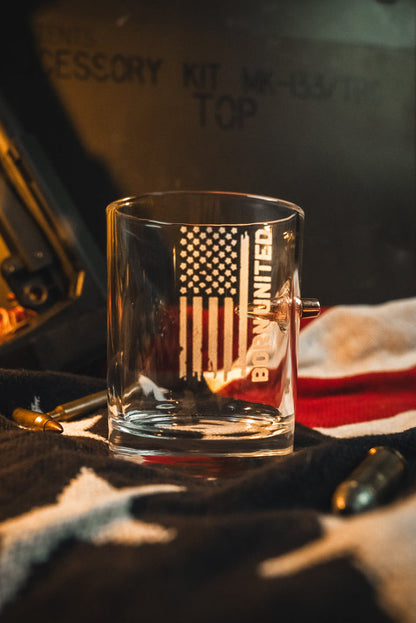 The Born United Bullet Whiskey Glass by Born United Private Label features an embedded bullet and American flag design, displayed on a folded flag with scattered bullets and military-style text in the dim background.