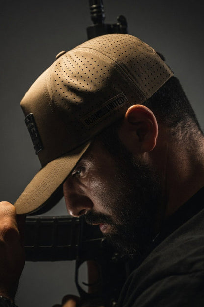 A bearded man wears the Born United Premium Camo Flag Snapback by Born United Private Label, holding the brim as he looks down. Dramatic lighting casts deep shadows on his face, with part of a rifle visible in the background.