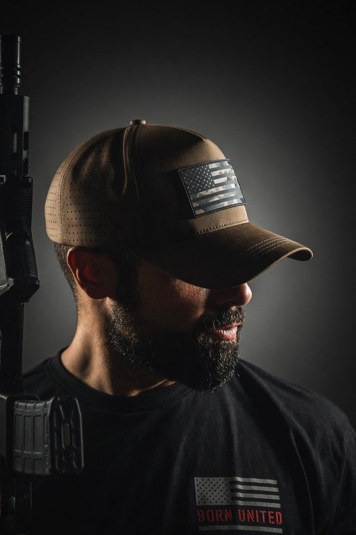 A bearded man in a Born United Premium Camo Flag Snapback and a black Born United Private Label t-shirt holds a rifle, standing in moody lighting against a dark background.