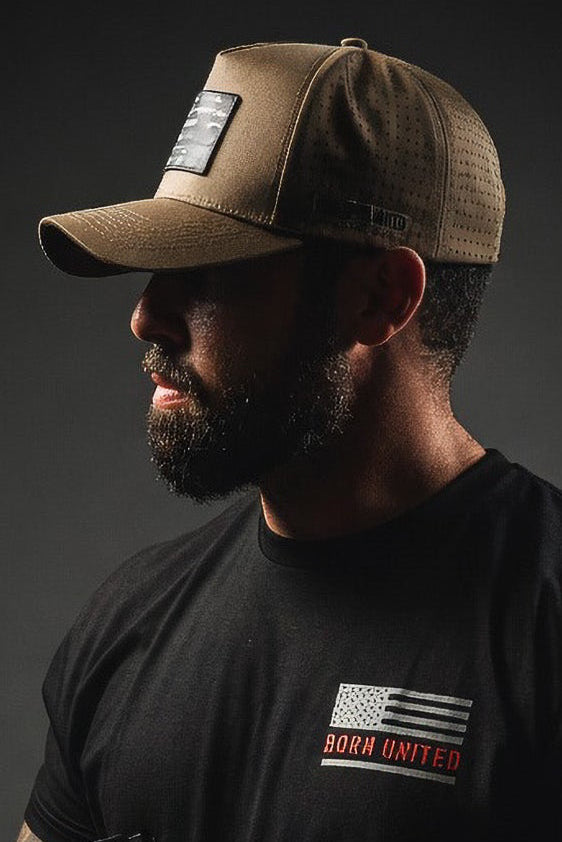 A bearded man in a brown cap wears the Born United Private Label Saints and Sinners OG Tee. The black shirt’s message stands out as he holds a firearm against a dark backdrop, his partially shadowed face turned to the side.