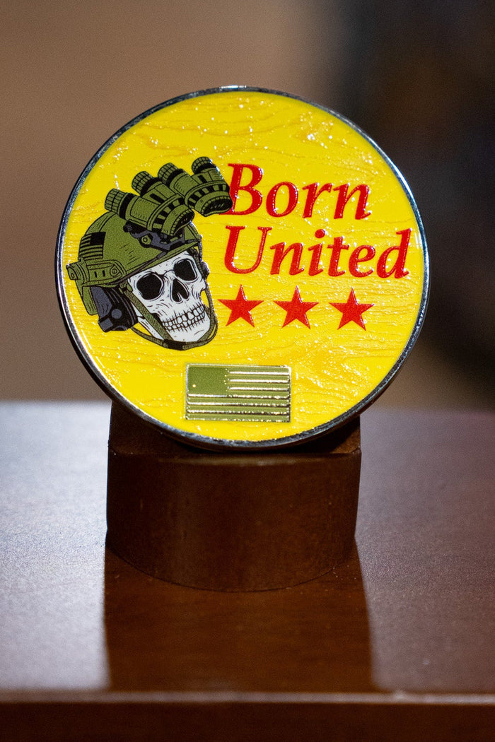 The Brass Pros Challenge Coin by Born United Private Label is a round yellow brass coin featuring a skull in a military helmet with night vision goggles, three red stars, and the words "Born United," displayed upright on a brown stand.