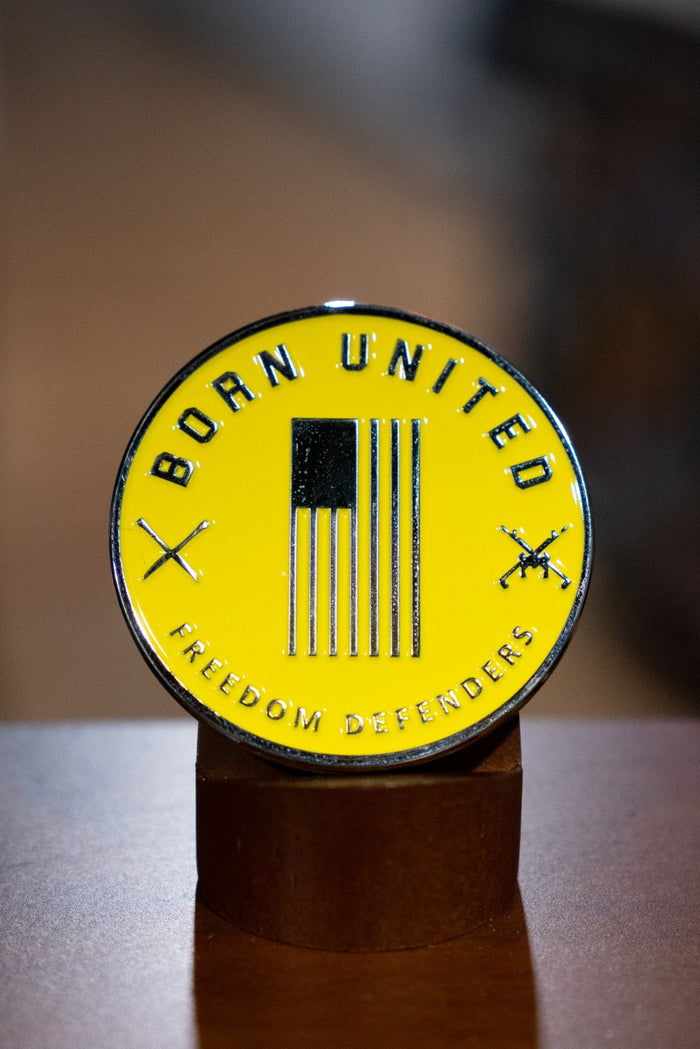 The Brass Pros Challenge Coin by Born United Private Label features a round yellow brass badge with a black vertical American flag, crossed rifles, and the words "BORN UNITED" and "FREEDOM DEFENDERS," displayed on a brown stand.