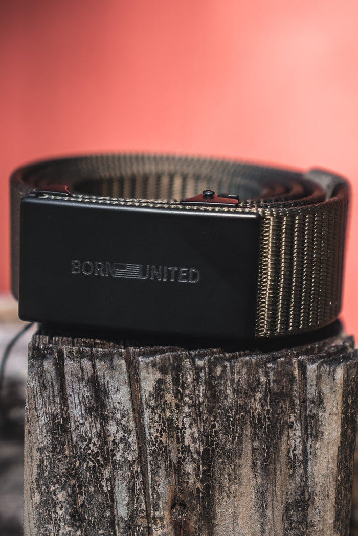 The BU Logo Belt - Army Green by Born United Private Label, featuring an adjustable design and a rectangular buckle engraved with BORN UNITED, is coiled on a weathered wooden post against a blurred reddish backdrop.