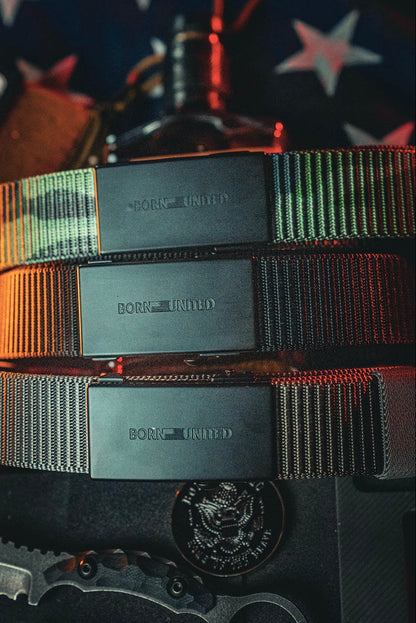 Three Born United Private Label BU Logo Belts in black, featuring adjustable fit and metal buckles with engraved logos, are stacked horizontally. An American flag and military gear appear partially in the background.