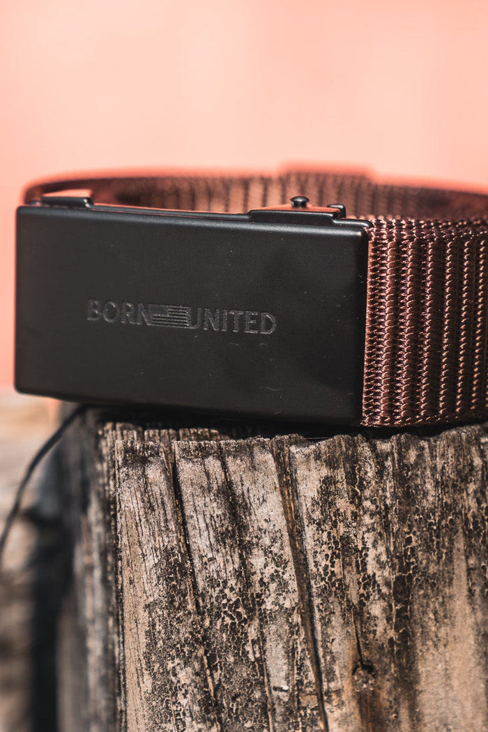 The BU Logo Belt - Brown by Born United Private Label features a brown woven design, black engraved buckle, and adjustable fit. Coiled on a rustic wooden post with a warm blurred background, it blends style with comfort.