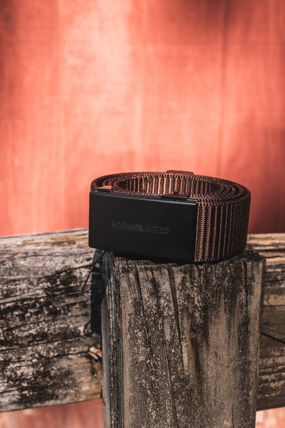The BU Logo Belt - Brown by Born United Private Label, featuring a black buckle, is displayed on a weathered wooden fence post with sunlight illuminating a reddish wooden wall in the background.
