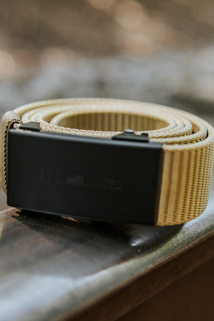 The BU Logo Belt - Coyote by Born United Private Label, featuring an adjustable tan strap and black buckle, is rolled up and positioned on a metal railway track with a blurred outdoor background of trees and sunlight.