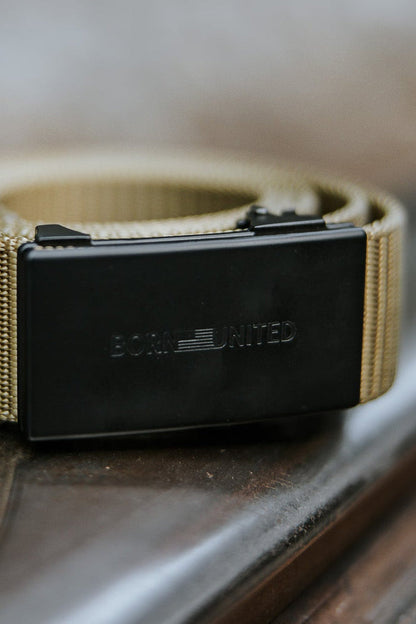 The BU Logo Belt - Coyote by Born United Private Label, featuring a black rectangular buckle engraved with "BORN UNITED," is displayed on a metal railing against a softly blurred background.