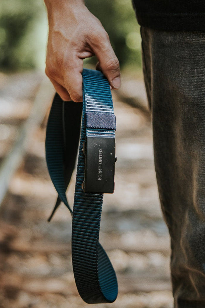 A person with a tattooed arm stands on railroad tracks, holding a Nikon camera strap. They're wearing jeans with the BU Logo Belt - Navy by Born United Private Label and a black shirt, outdoors with green scenery in the background.