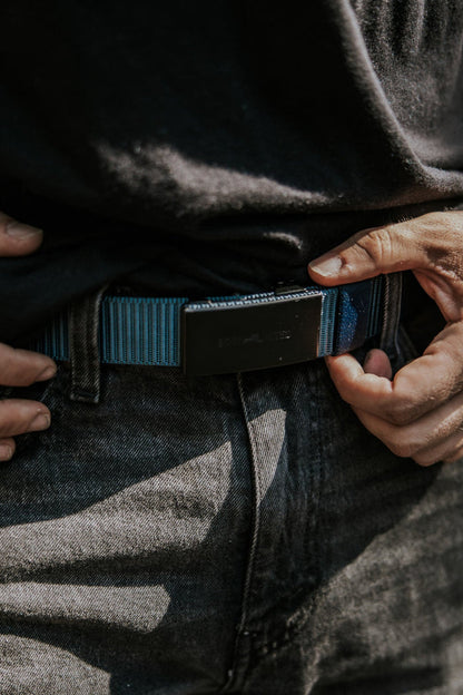 A person wearing a black shirt and dark jeans adjusts the BU Logo Belt - Navy by Born United Private Label, focusing on their hands and waist in natural outdoor lighting.