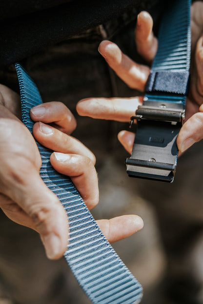 Close-up of hands fastening the BU Logo Belt - Navy from Born United Private Label, featuring an adjustable blue and gray striped nylon strap with a black metal buckle. The blurred background emphasizes the details of the belt and hands.