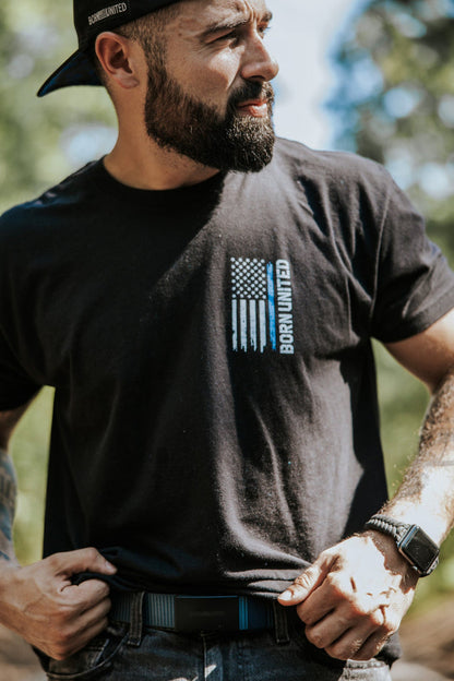 A bearded man outdoors in sunlight, wearing a black “Born United” tee with an American flag print, adjusts his Born United Private Label BU Logo Belt - Navy. He has tattooed arms, a smartwatch, and wears his black cap backwards.