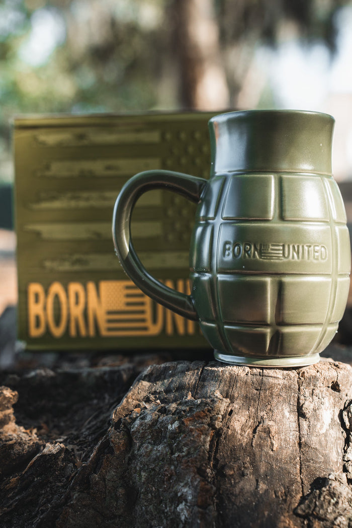 The BU Sip Sip Boom Mug by Born United Private Label—a green, grenade-shaped coffee mug embossed with BORN UNITED—rests on a tree stump; its matching American flag box with BORN UNITED text is blurred in the background.