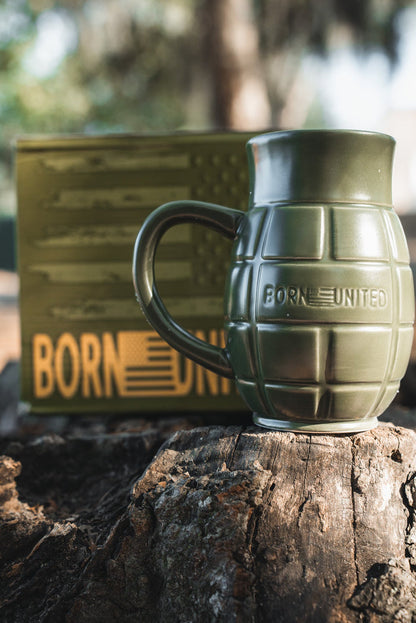 The BU Sip Sip Boom Mug by Born United Private Label—a green, grenade-shaped coffee mug embossed with BORN UNITED—rests on a tree stump; its matching American flag box with BORN UNITED text is blurred in the background.
