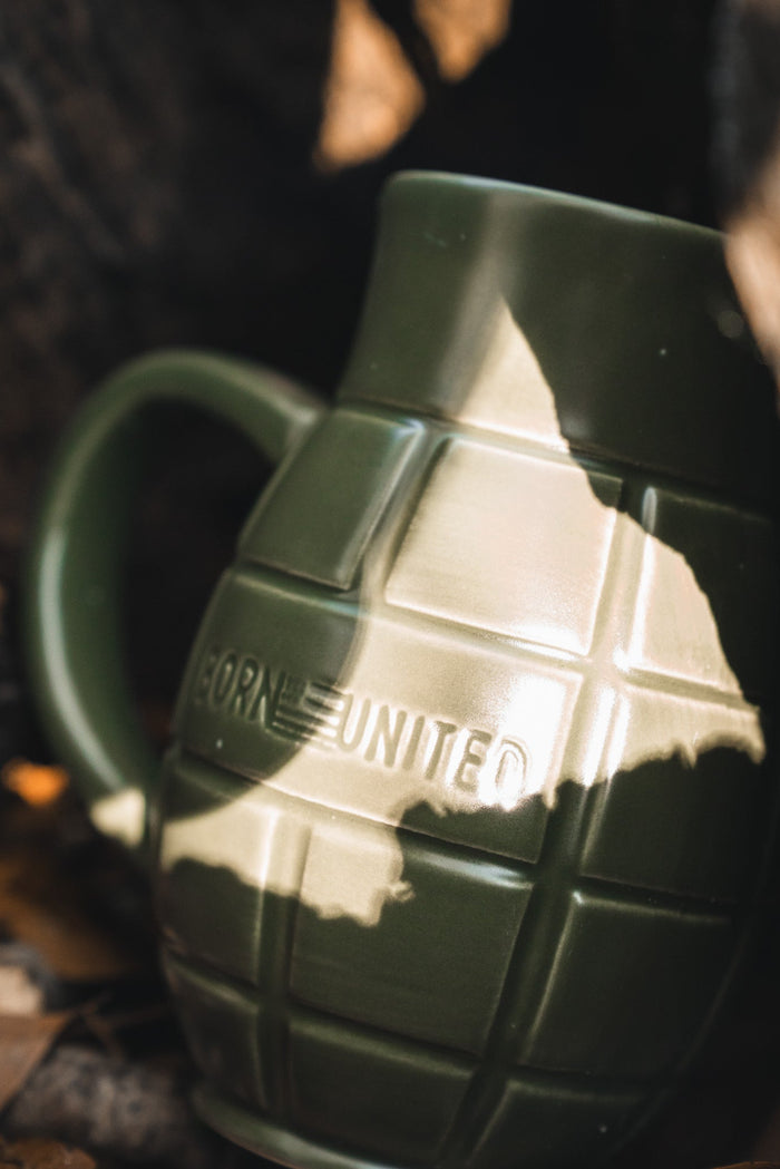 The BU Sip Sip Boom Mug by Born United Private Label is a hand-crafted green mug embossed with "BORN UNITED," catching the sunlight among blurred natural surroundings—a bold conversation starter for any setting.