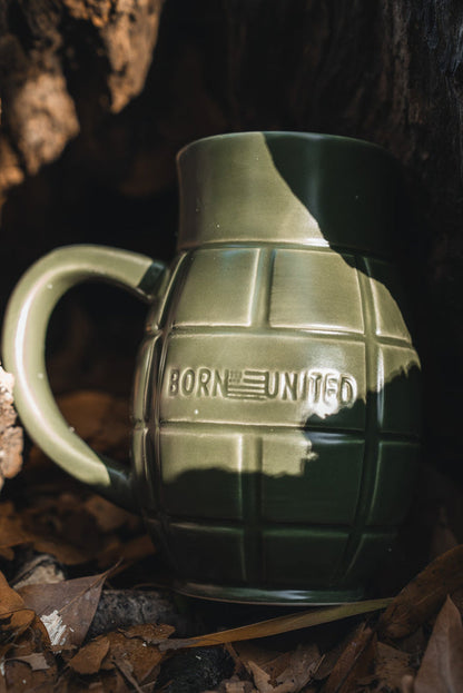 The BU Sip Sip Boom Mug by Born United Private Label is a green, hand-crafted grenade mug featuring "BORN UNITED" and an American flag—an eye-catching conversation starter, shown here among brown leaves and partial shadow.