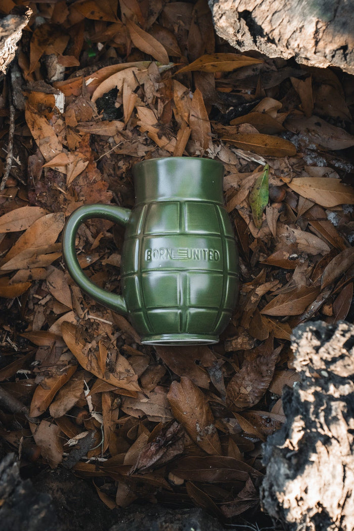 The BU Sip Sip Boom Mug by Born United Private Label, featuring BORN UNITED embossing, rests on dry brown leaves amid pieces of tree bark.