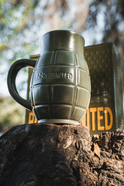 The BU Sip Sip Boom Mug by Born United Private Label, shaped like a hand grenade and embossed with "BORN UNITED," rests on a tree stump outdoors with its matching box blurred in the background—a unique addition to any collection.