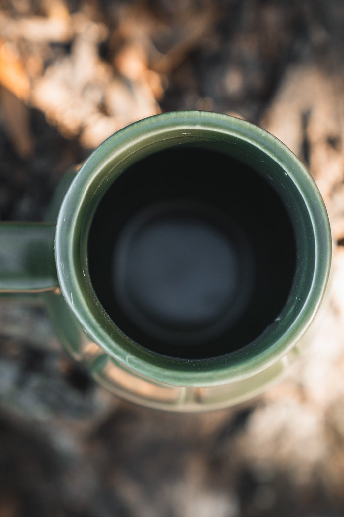 Close-up, top-down view of the BU Sip Sip Boom Mug by Born United Private Label, hand-crafted with a handle and placed on a blurred outdoor surface, possibly soil or wood.
